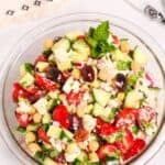 A glass bowl filled with a vibrant Chickpea Feta Salad—chopped cucumbers, tomatoes, olives, red onions, and fresh mint—sits on a white surface.