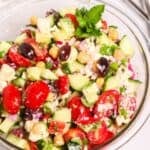 A glass bowl of Chickpea Feta Salad with cherry tomatoes, cucumber, olives, red onion, and fresh herbs. Two forks rest beside the bowl on a white surface.
