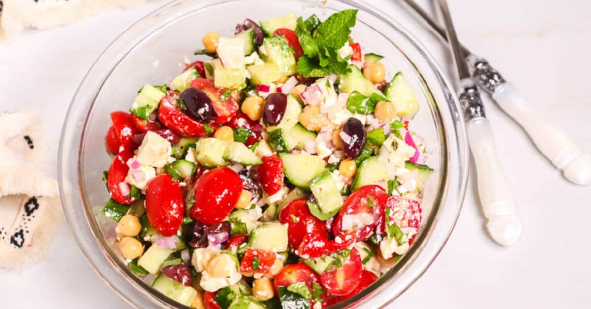 A glass bowl of Chickpea Feta Salad with cherry tomatoes, cucumber, olives, red onion, and fresh herbs. Two forks rest beside the bowl on a white surface.