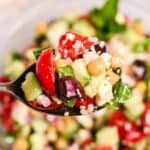 A spoonful with chopped cucumber, tomato, red onion, olives, parsley, and feta cheese held above a bowl of the same vibrant salad.