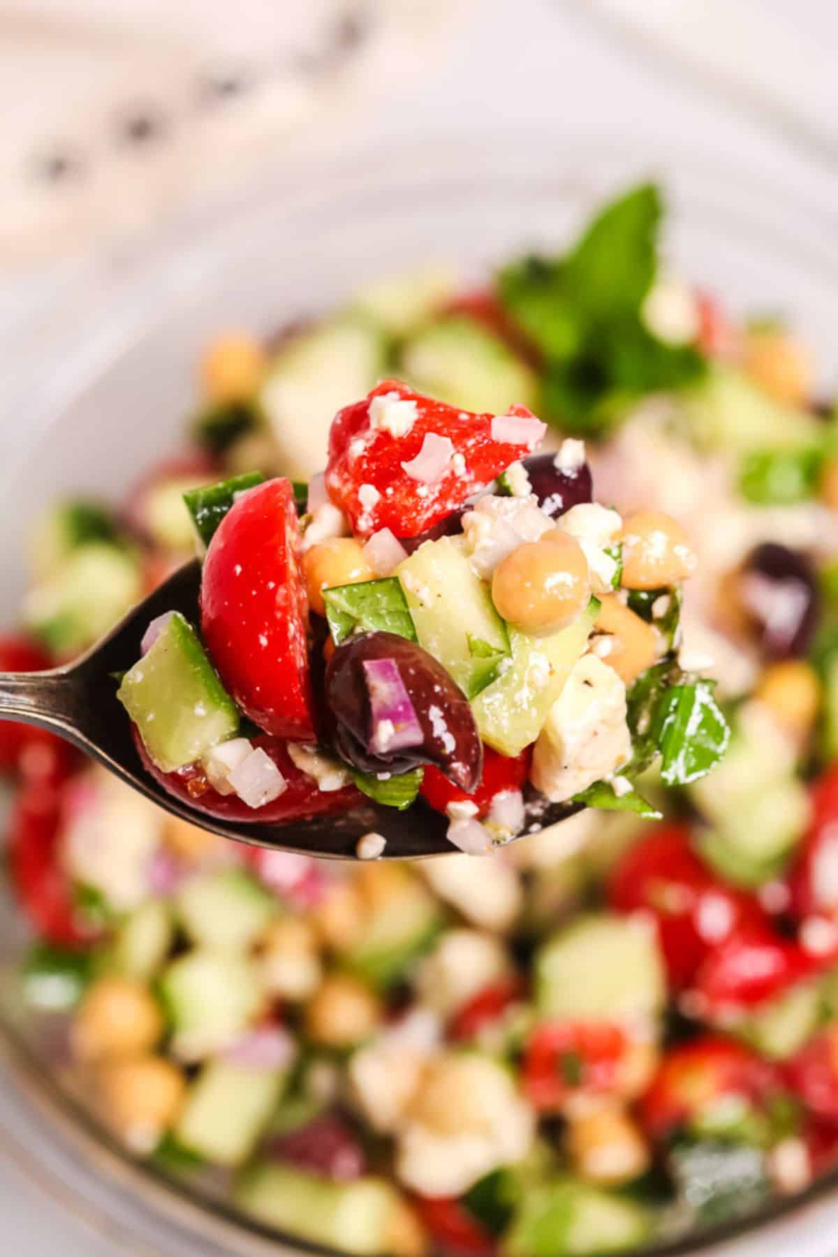 A spoonful with chopped cucumber, tomato, red onion, olives, parsley, and feta cheese held above a bowl of the same vibrant salad.