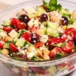 A glass bowl featuring tomatoes, cucumbers, red onion, black olives, fresh herbs, and creamy feta cheese.