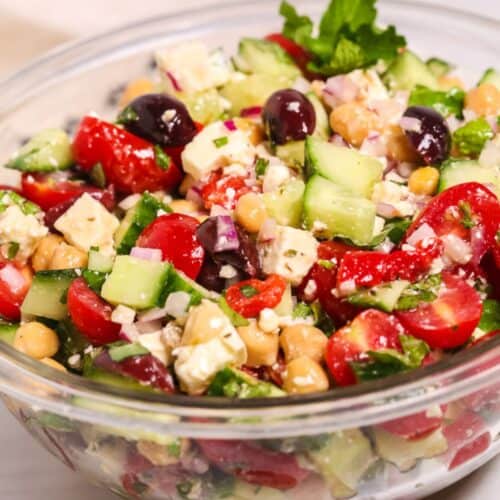 A glass bowl featuring tomatoes, cucumbers, red onion, black olives, fresh herbs, and creamy feta cheese.