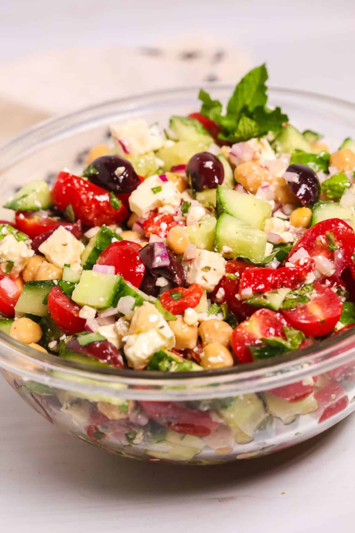 A glass bowl  featuring tomatoes, cucumbers, red onion, black olives, fresh herbs, and creamy feta cheese.