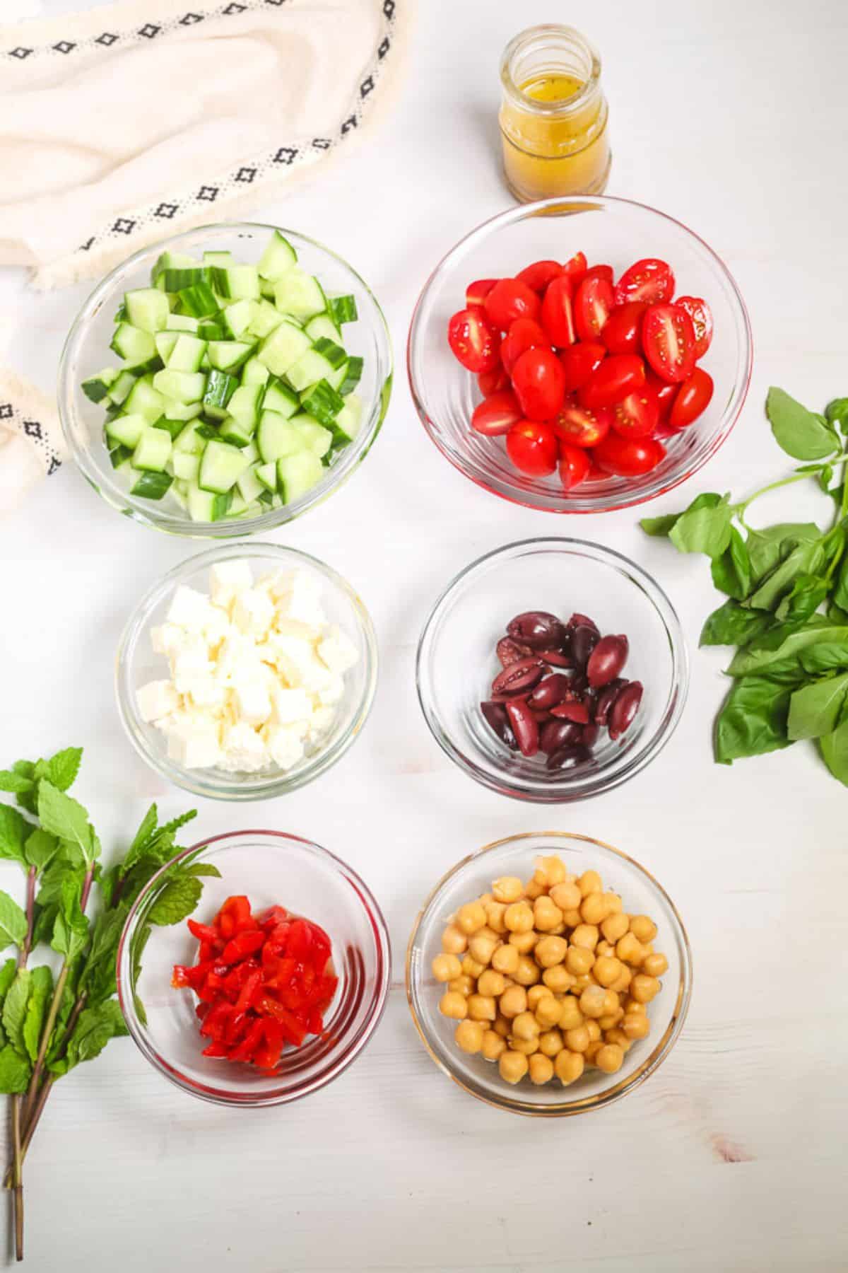 Seven bowls contain ingredients chopped cucumber, grape tomatoes, feta cheese, Kalamata olives, roasted red peppers, and chickpeas. A jar of dressing and fresh herbs are arranged nearby on a white surface.