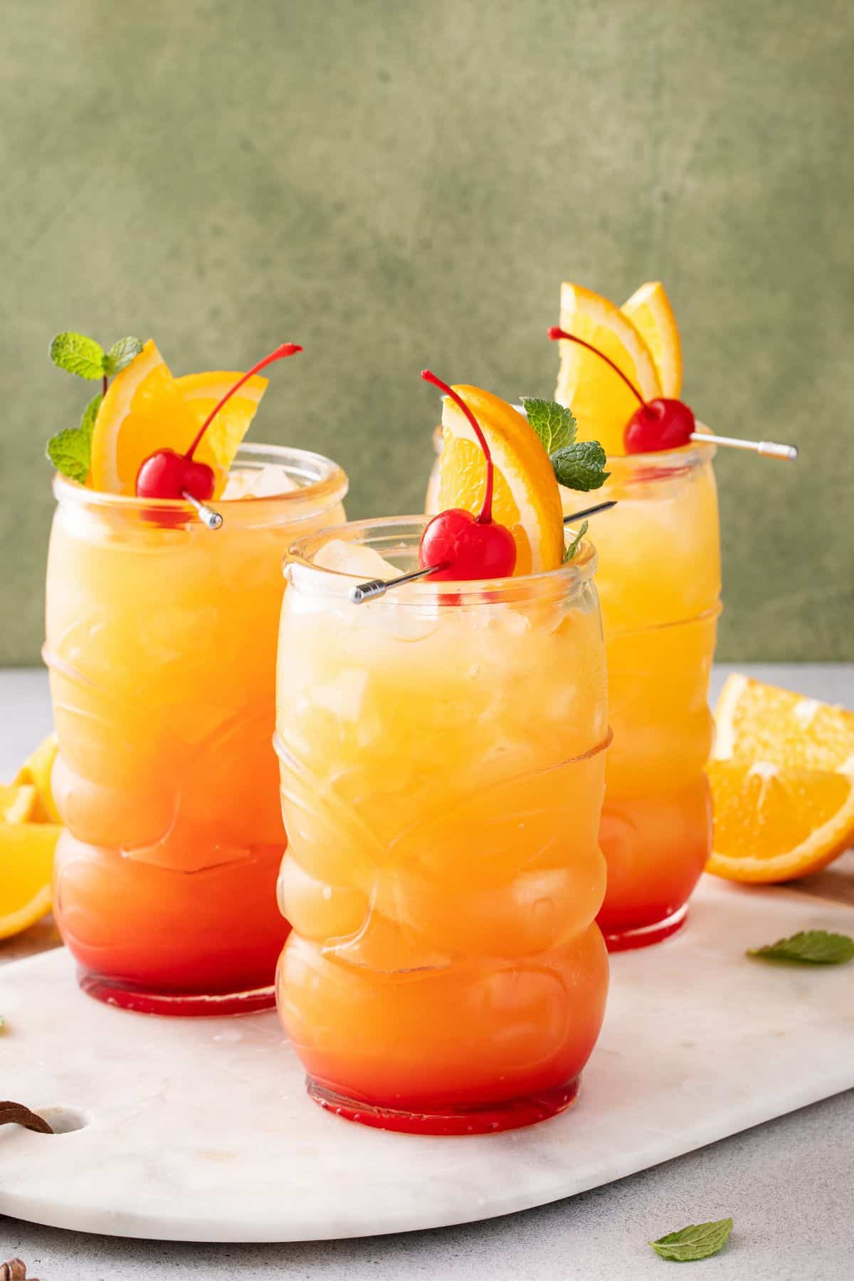 Three tiki glasses filled with orange and red layered Jamaican rum punch cocktails, garnished with orange slices, cherries, and mint, are placed on a marble surface with orange wedges nearby.