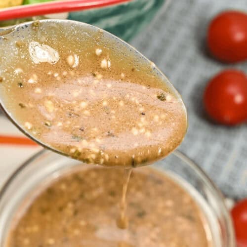 A spoon holding a brown, textured creamy balsamic dressing is being poured into a glass bowl with more of the same dressing. A salad with lettuce and tomatoes is in the background.