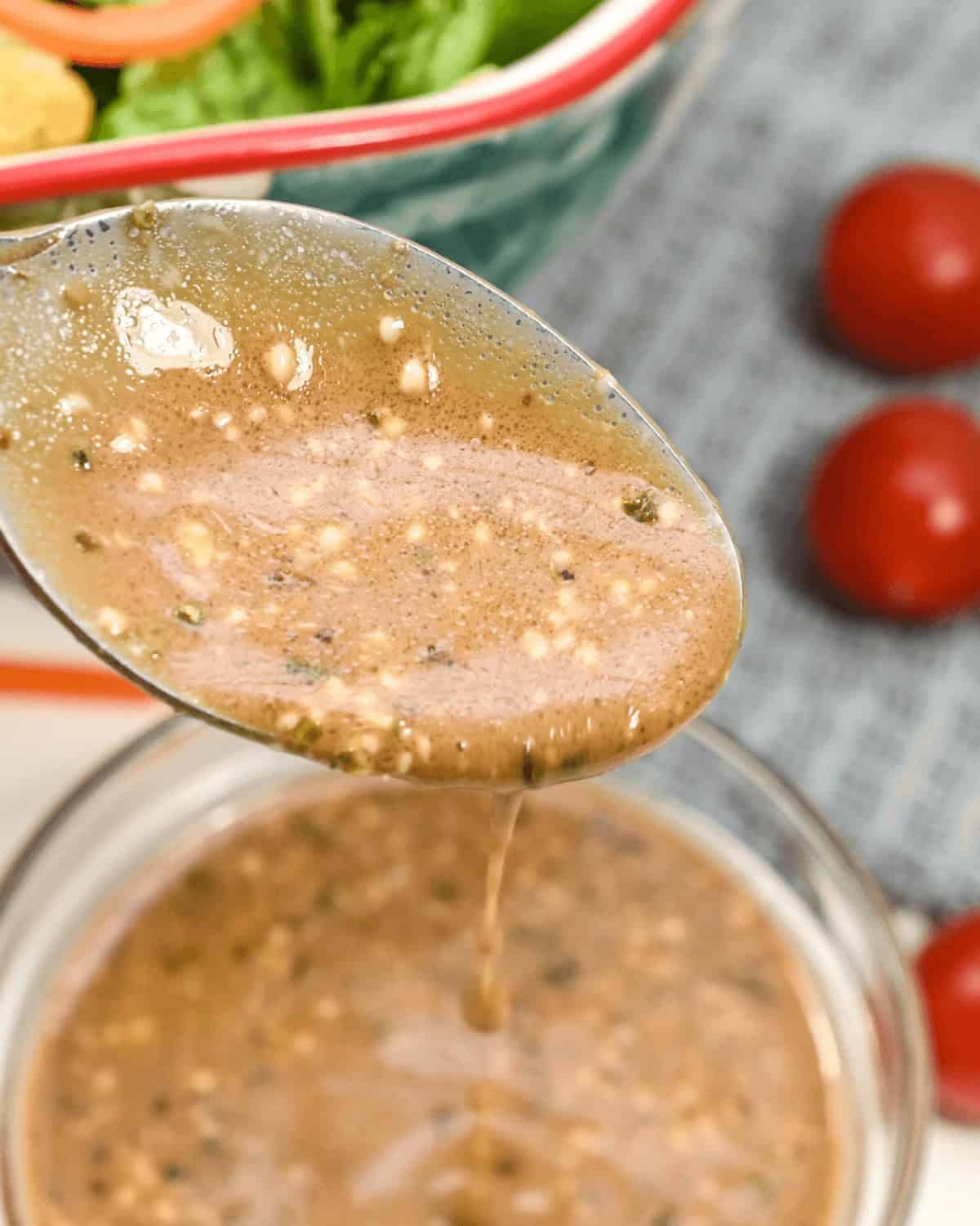 A spoon holding a brown, textured creamy balsamic dressing is being poured into a glass bowl with more of the same dressing. A salad with lettuce and tomatoes is in the background.