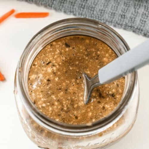 A jar filled with a brown, speckled liquid mixture, resembling creamy balsamic dressing, with a spoon standing inside it. The jar is placed on a white surface with a textured gray fabric in the background.