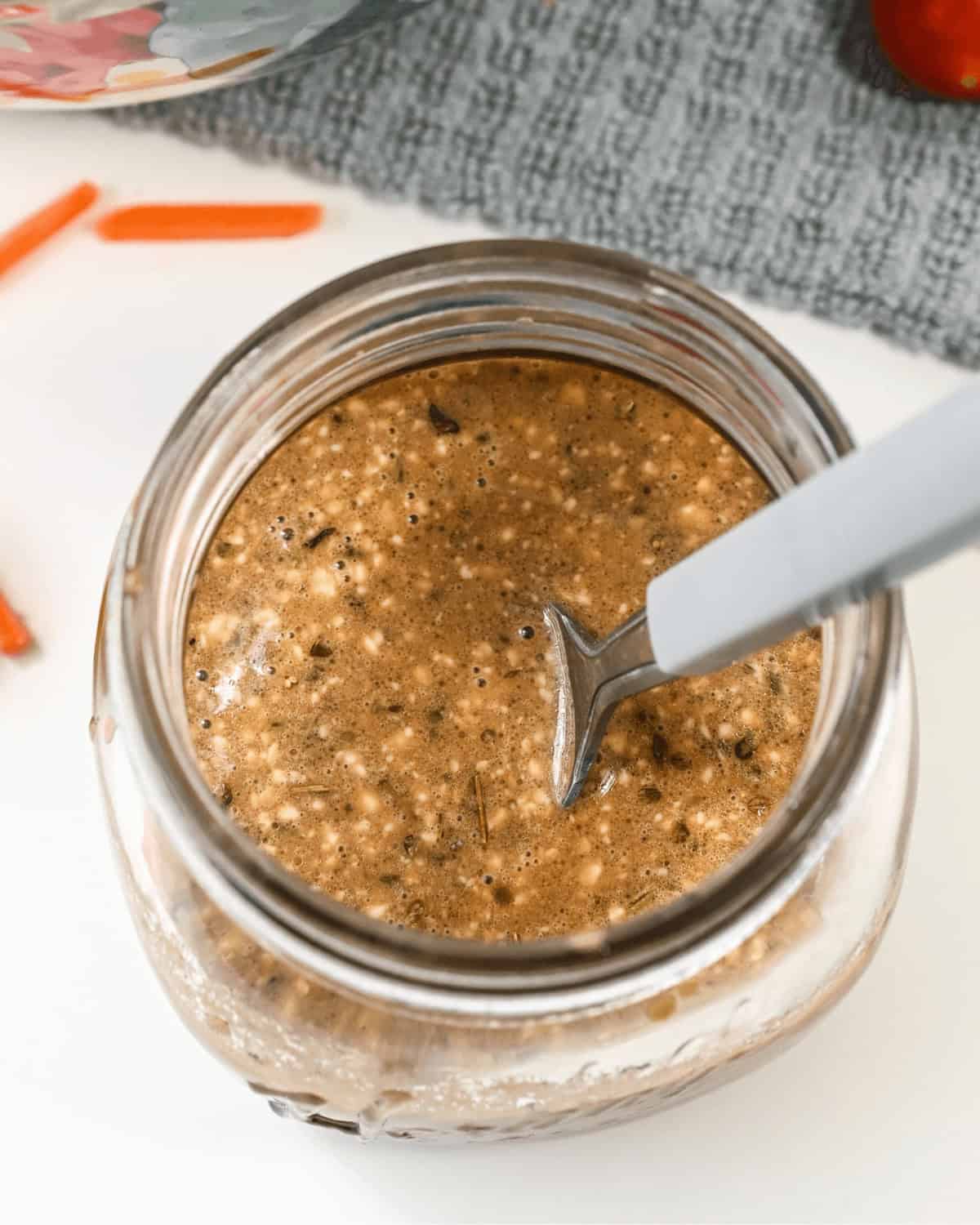 A jar filled with a brown, speckled liquid mixture, resembling creamy balsamic dressing, with a spoon standing inside it. The jar is placed on a white surface with a textured gray fabric in the background.