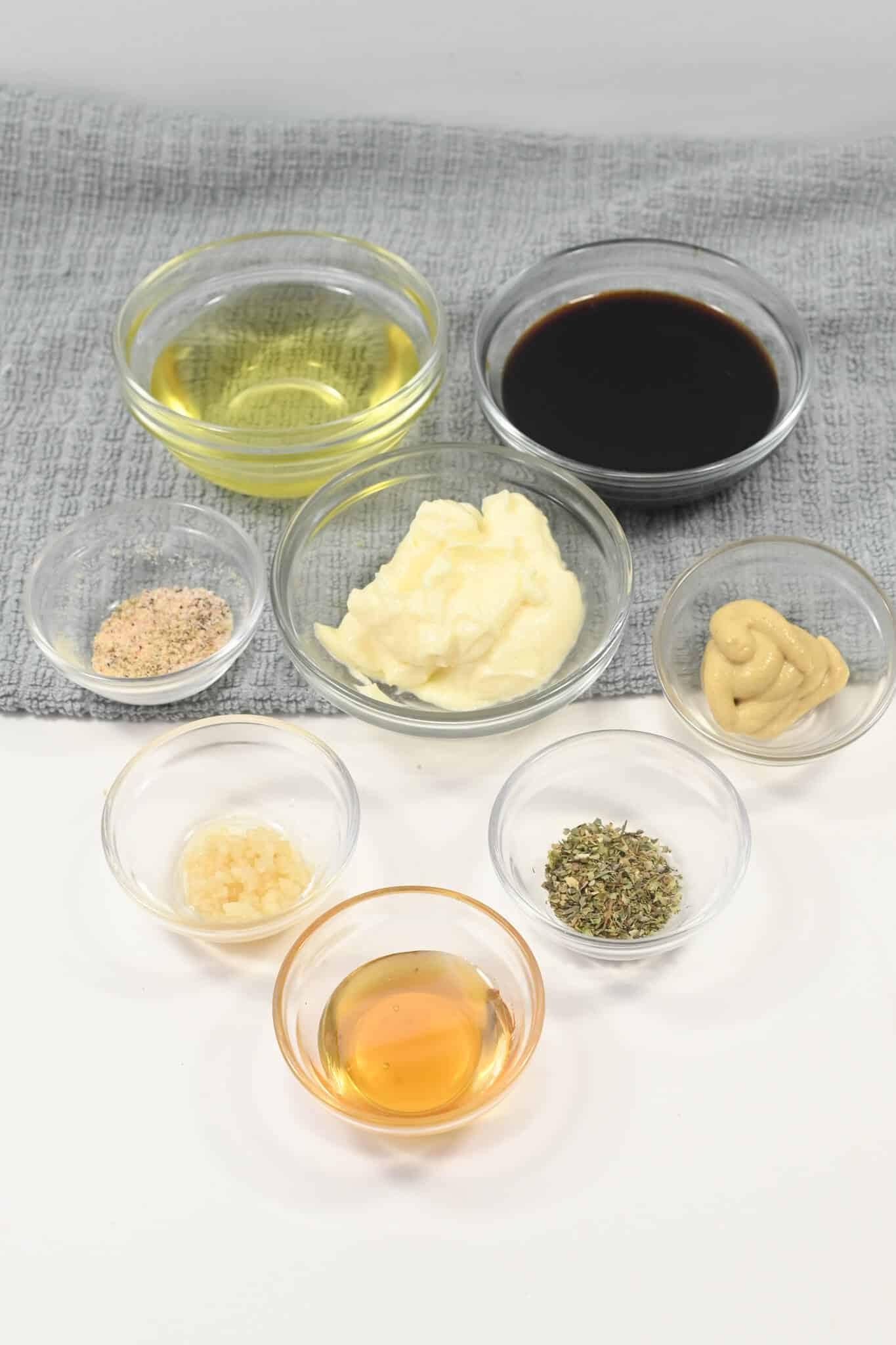 A collection of various ingredients in small glass bowls, including oil, soy sauce, mayonnaise, creamy balsamic dressing, mustard, seasoning, minced garlic, dried herbs, and a light brown liquid, arranged on a gray cloth.