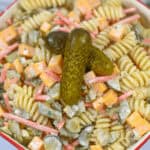 A bowl of creamy dill pickle noodle salad with chunks of cheese, diced pickles, and herbs, garnished with a whole pickle on top.