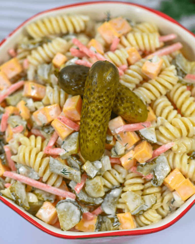 A bowl of creamy dill pickle noodle salad with chunks of cheese, diced pickles, and herbs, garnished with a whole pickle on top.