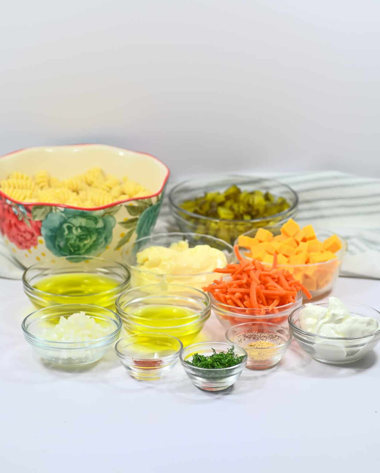 Various ingredients for cooking arranged in small bowls, including pasta, cheese, vegetables, dill pickle, and spices, on a neutral background.