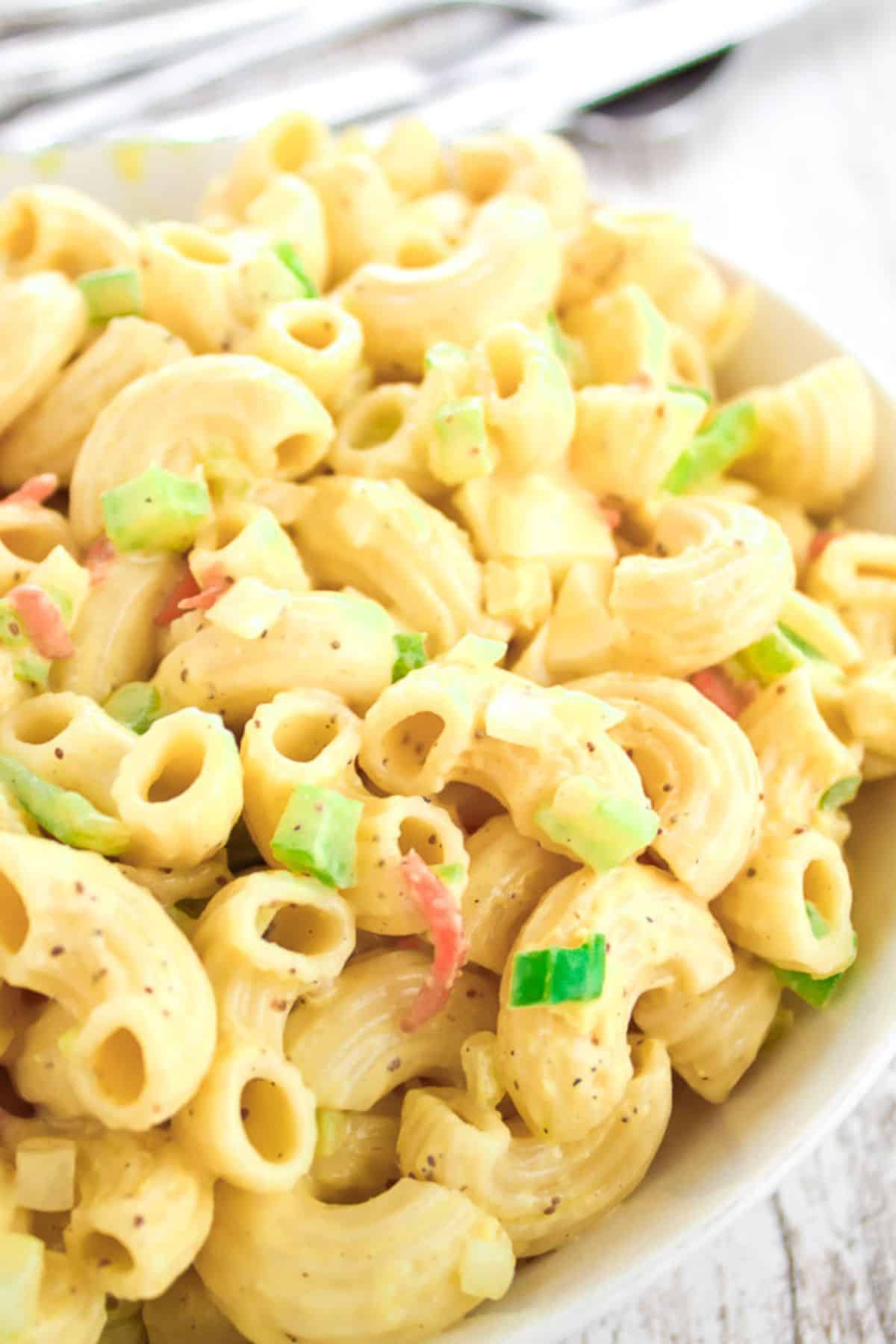A close-up of this Easy Macaroni Salad Recipe shows elbow pasta, chopped celery, and peppers in a creamy dressing, served in a white bowl.
