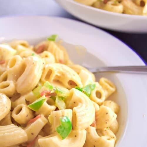 Close-up of an Easy Macaroni Salad Recipe, showcasing creamy macaroni mixed with chopped green bell peppers and red pieces, with a bowl of extra salad in the background.