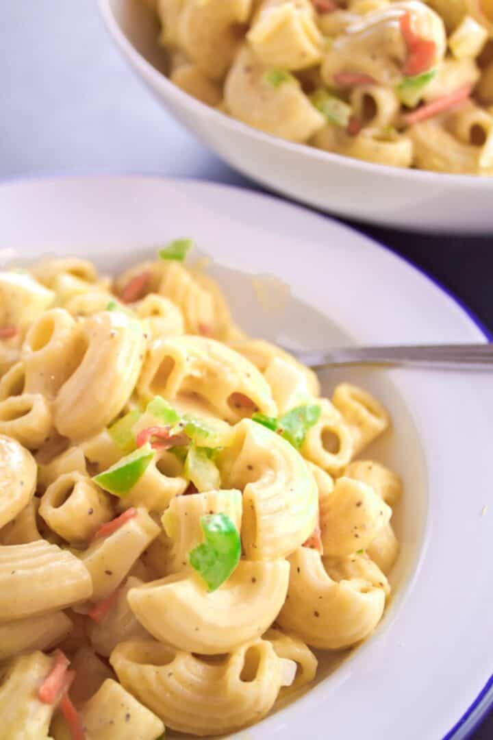 Close-up of an Easy Macaroni Salad Recipe, showcasing creamy macaroni mixed with chopped green bell peppers and red pieces, with a bowl of extra salad in the background.