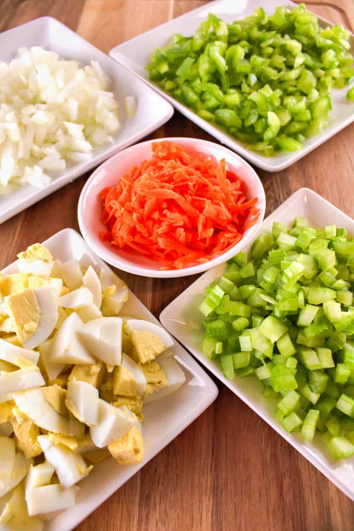 Five plates on a wooden surface hold chopped hard-boiled eggs, celery, onions, green bell peppers, and a central dish of shredded carrots.