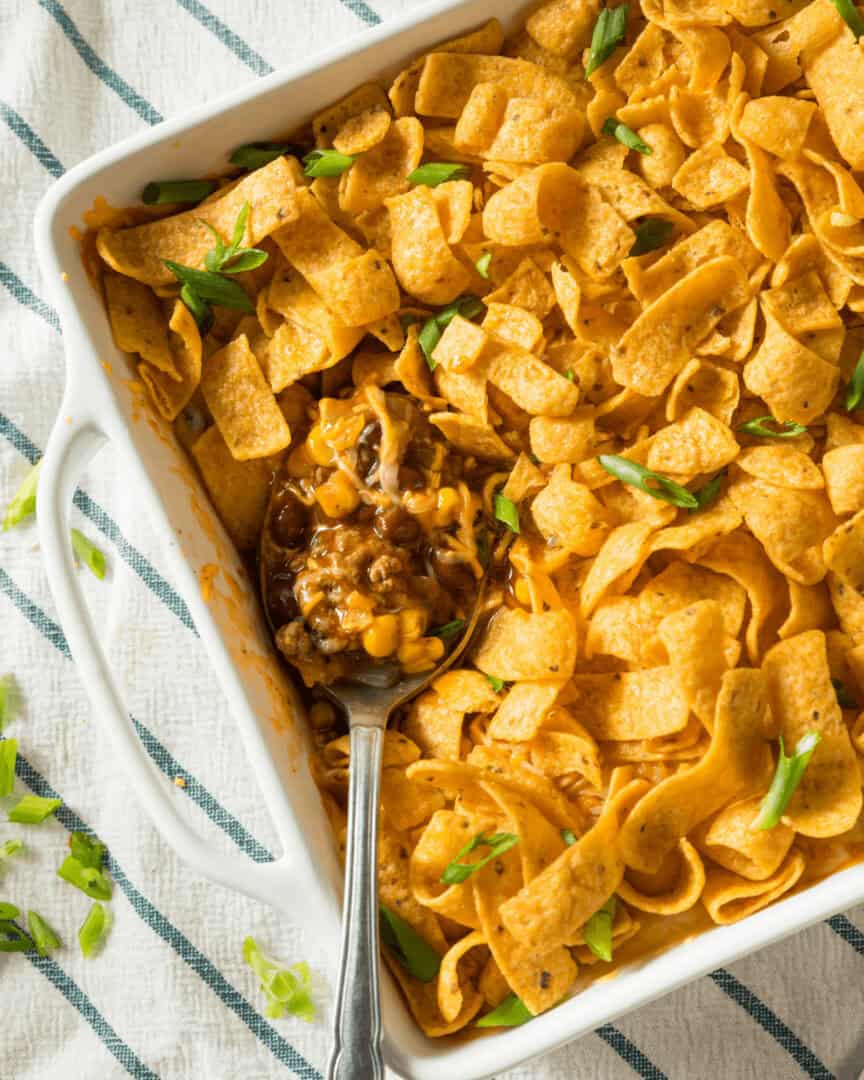 A casserole dish filled with Frito Chili Pie, topped with corn chips, melted cheese, and garnished with green onions. A spoon is scooping out a serving.
