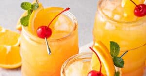 Three glasses of orange cocktails with ice, each garnished with an orange slice, a cherry, and a sprig of mint—perfect for enjoying a refreshing Jamaican rum punch.