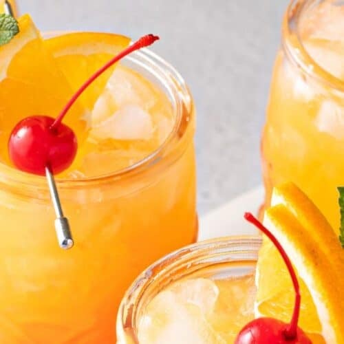 Three glasses of orange cocktails with ice, each garnished with an orange slice, a cherry, and a sprig of mint—perfect for enjoying a refreshing Jamaican rum punch.