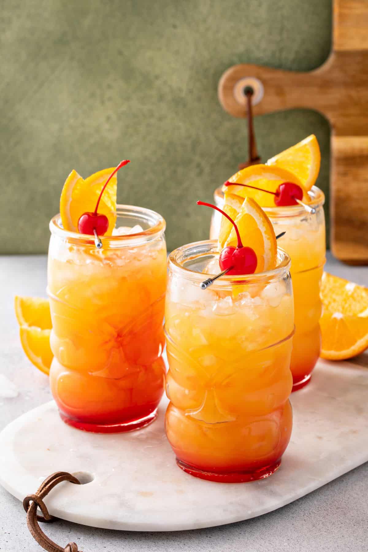 Three tropical cocktails with orange slices and cherries, served in tall glasses with ice on a white marble board—perfect for savoring the vibrant flavors of Jamaican rum punch.