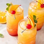 Three glasses of a Jamaican rum punch cocktail garnished with orange slices, maraschino cherries, and mint leaves, served on a white surface with extra orange slices in the background.