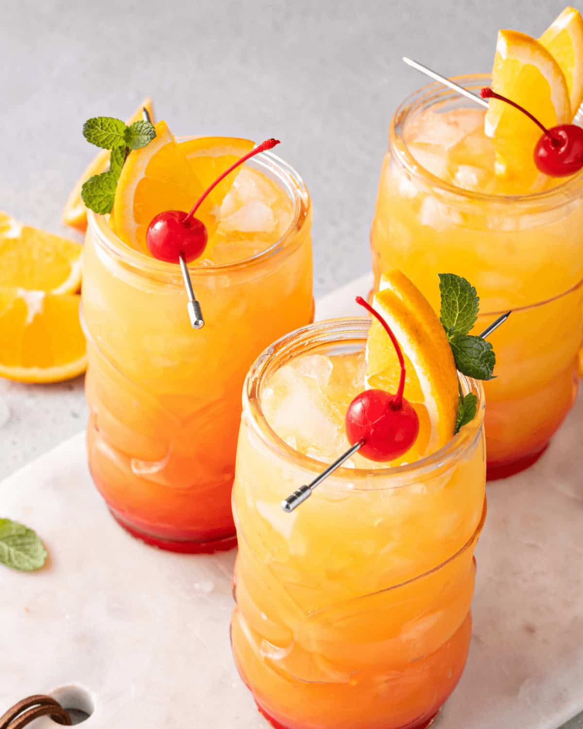 Three glasses of a Jamaican rum punch cocktail garnished with orange slices, maraschino cherries, and mint leaves, served on a white surface with extra orange slices in the background.