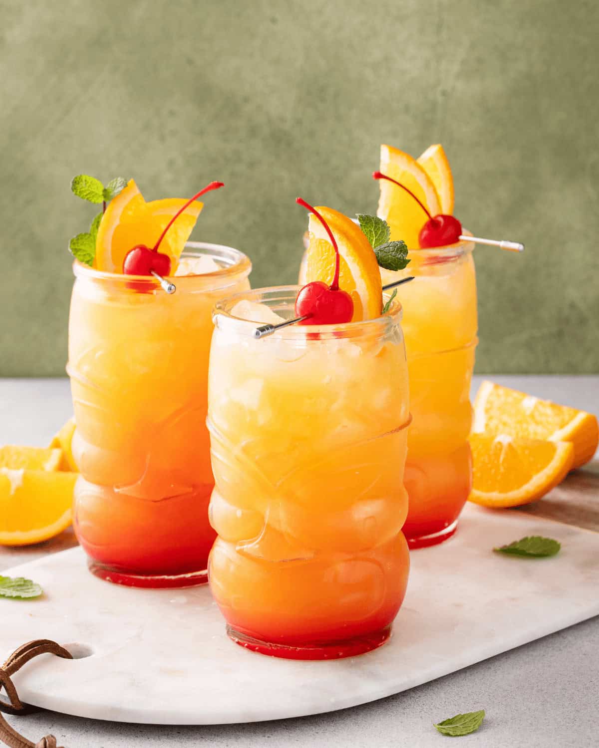 Three tropical cocktails featuring Jamaican rum punch, garnished with orange slices and cherries, served over ice in textured glasses, positioned on a marble surface with orange wedges and mint leaves.