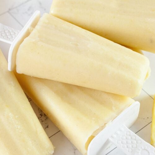 Four creamy yellow Pineapple Popsicles with white handles are stacked on a light surface, with a piece of pineapple visible in the background.