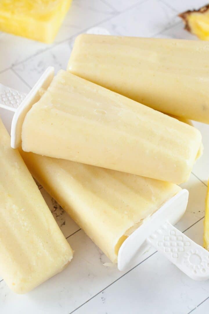 Four creamy yellow Pineapple Popsicles with white handles are stacked on a light surface, with a piece of pineapple visible in the background.