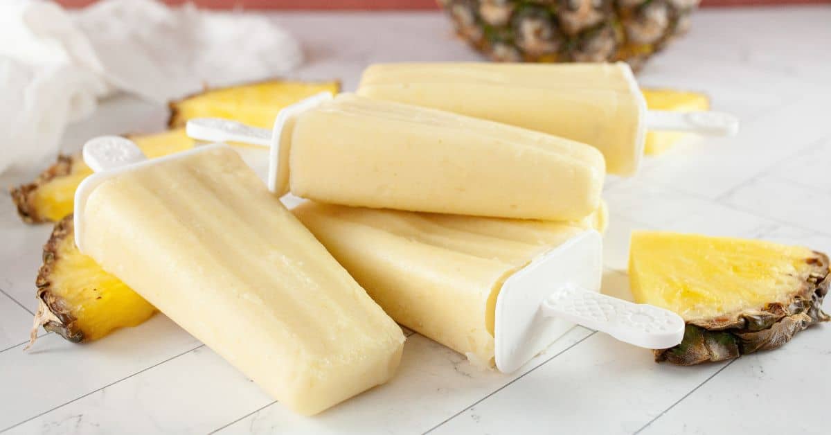 Four creamy yellow Pineapple Popsicles are stacked on a white surface with pineapple slices and a fresh pineapple in the background.