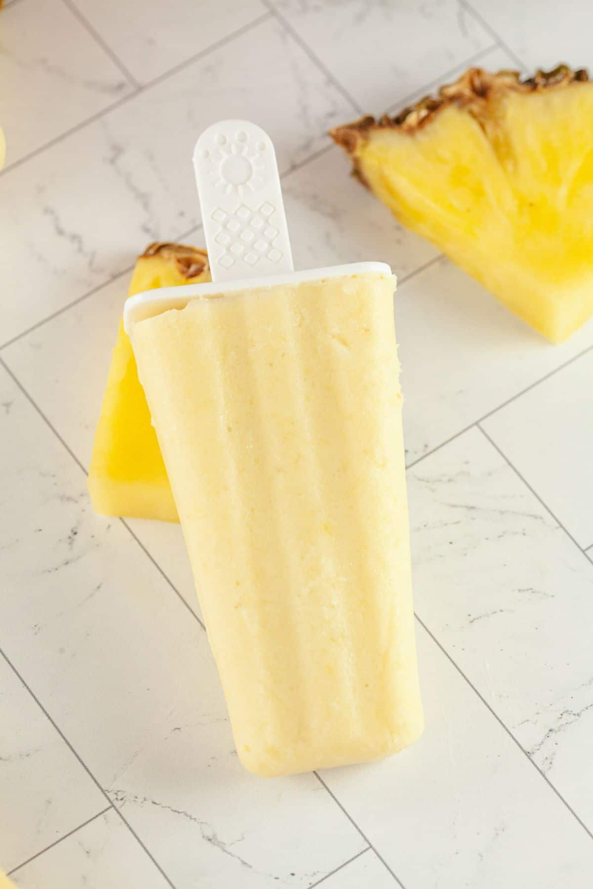 A yellow Pineapple Popsicle with a white handle is placed on a white tiled surface, with pineapple slices in the background.