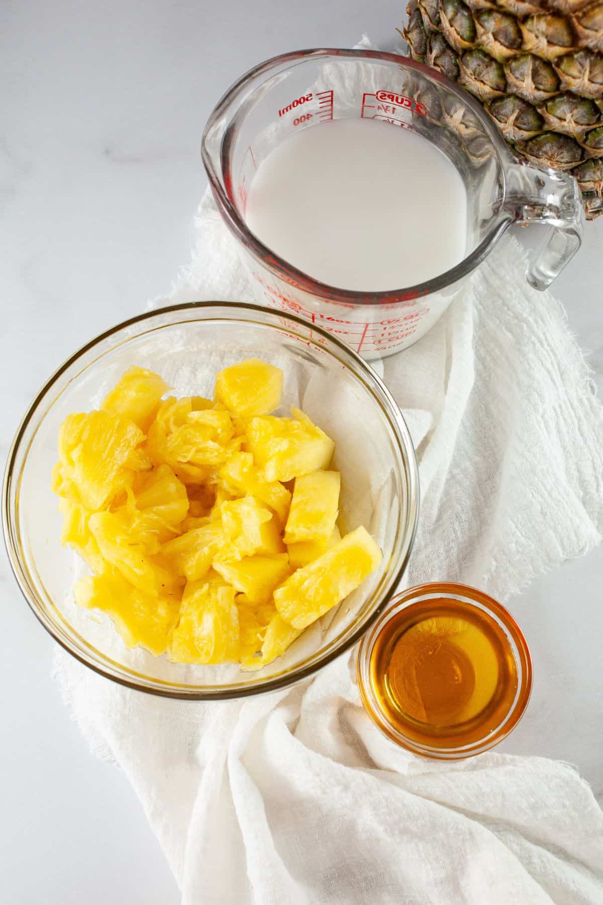 A glass bowl of pineapple chunks, a measuring cup of coconut milk, and a dish of honey are arranged on a white cloth with a partial pineapple.