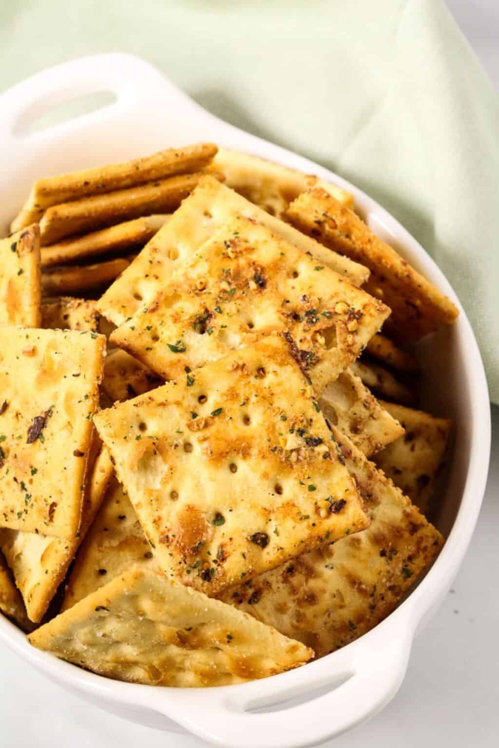 A white dish filled with seasoned, square crackers from a spicy crackers recipe, with a light green cloth in the background.