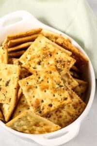 A white dish filled with seasoned, square crackers from a spicy crackers recipe, with a light green cloth in the background.