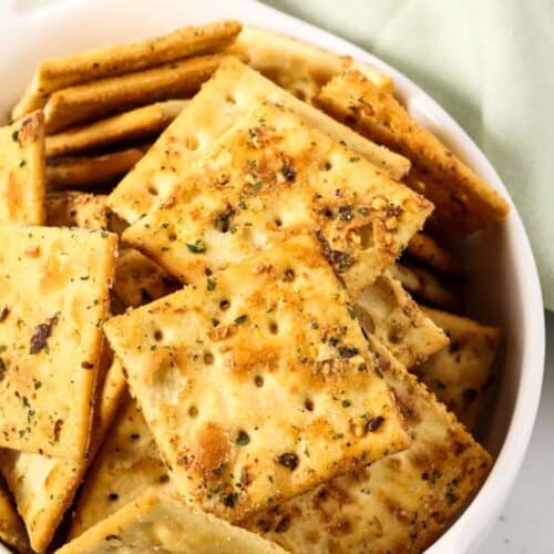 A white dish filled with seasoned, square crackers from a spicy crackers recipe, with a light green cloth in the background.
