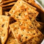 A bowl filled with seasoned square crackers, topped with visible herbs and spices, perfect for trying out your favorite Spicy Crackers Recipe.