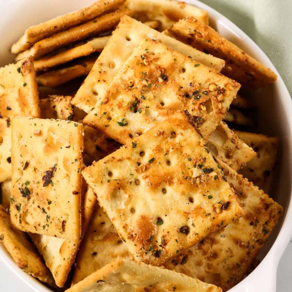 A bowl filled with seasoned square crackers topped with herbs and spices, perfect for trying out your favorite Spicy Crackers Recipe.