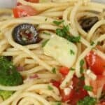 A close-up of Spaghetti Pasta Salad mixed with broccoli, black olives, diced tomatoes, parsley, and chunks of cheese in a white bowl.