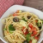 A bowl of Spaghetti Pasta Salad with broccoli, black olives, diced tomatoes, cucumber, and chopped parsley sits on a wooden surface beside a red checkered cloth.