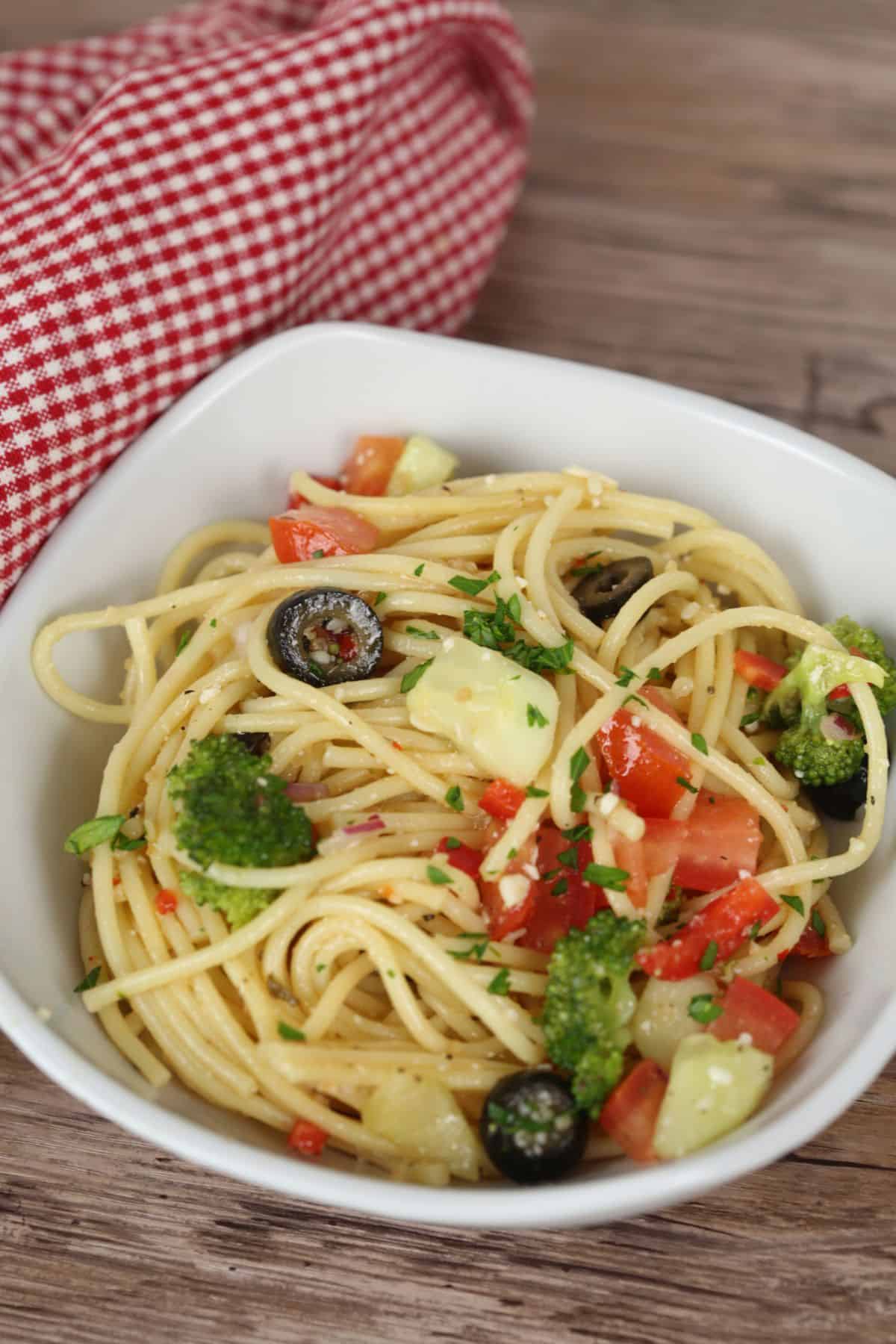 A bowl of Spaghetti Pasta Salad with broccoli, black olives, diced tomatoes, cucumber, and chopped parsley sits on a wooden surface beside a red checkered cloth.