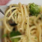 A close-up of a fork holding spaghetti pasta salad with broccoli, with more pasta visible in a white bowl below.