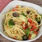 A bowl of Spaghetti Pasta Salad with chopped tomatoes, broccoli, black olives, cucumber, red onion, and parsley sits on a wooden surface.