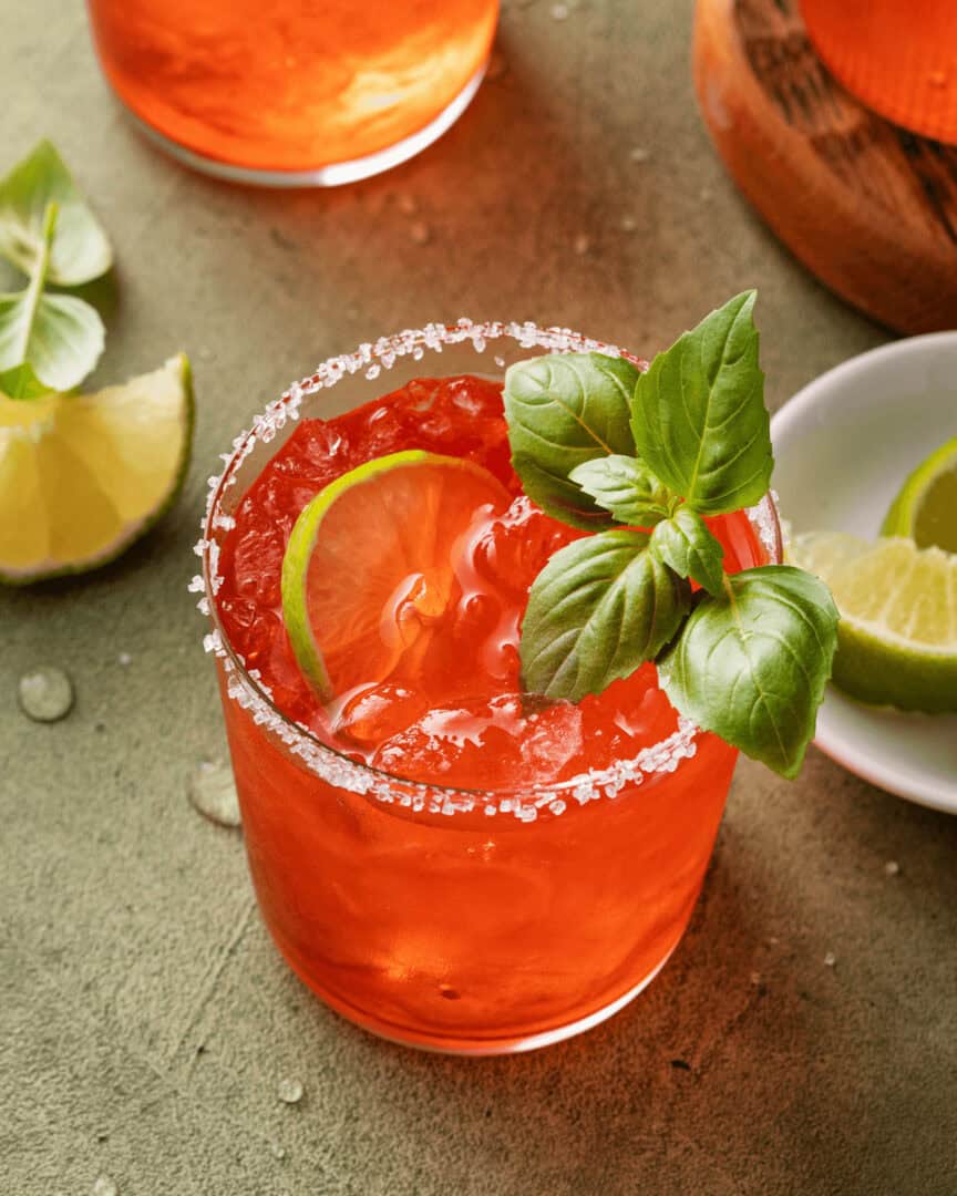 A glass with an strawberry basil margarita garnished with a lime slice, basil leaves, and a salted rim. A wedge of lime and basil leaves are on the surface, with a white plate of lime wedges in the background.