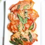 A white rectangular plate of prosciutto-wrapped Veal Saltimbocca, garnished with sage leaves and a light sauce.