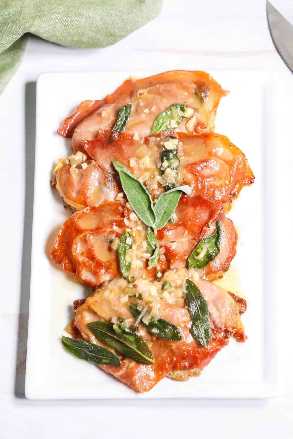 A white rectangular plate of prosciutto-wrapped Veal Saltimbocca, garnished with sage leaves and a light sauce.
