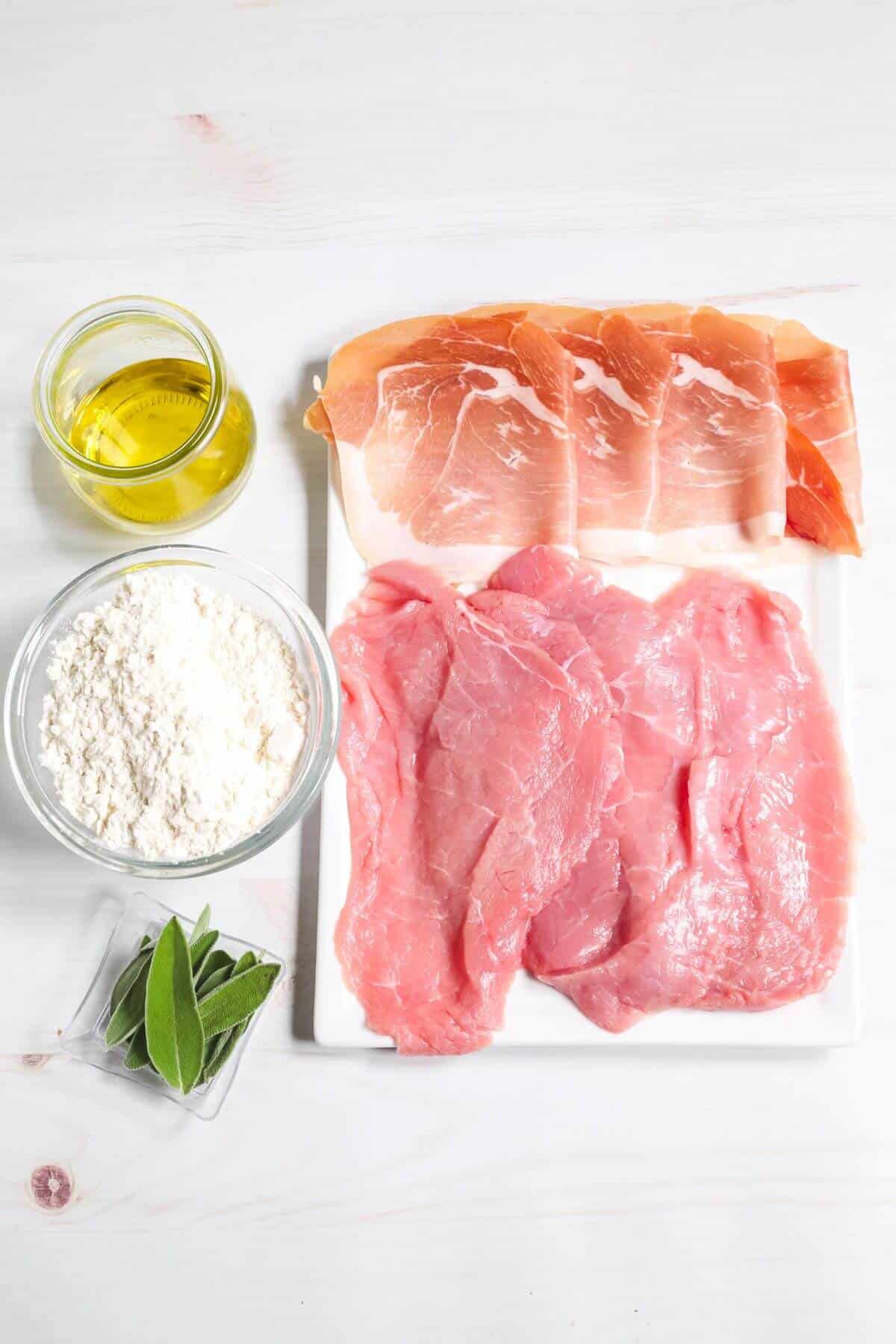 A preparation for Veal Saltimbocca: raw veal cutlets, prosciutto slices, a bowl of flour, a glass jug of olive oil, and sprigs of fresh sage are arranged on a white surface.