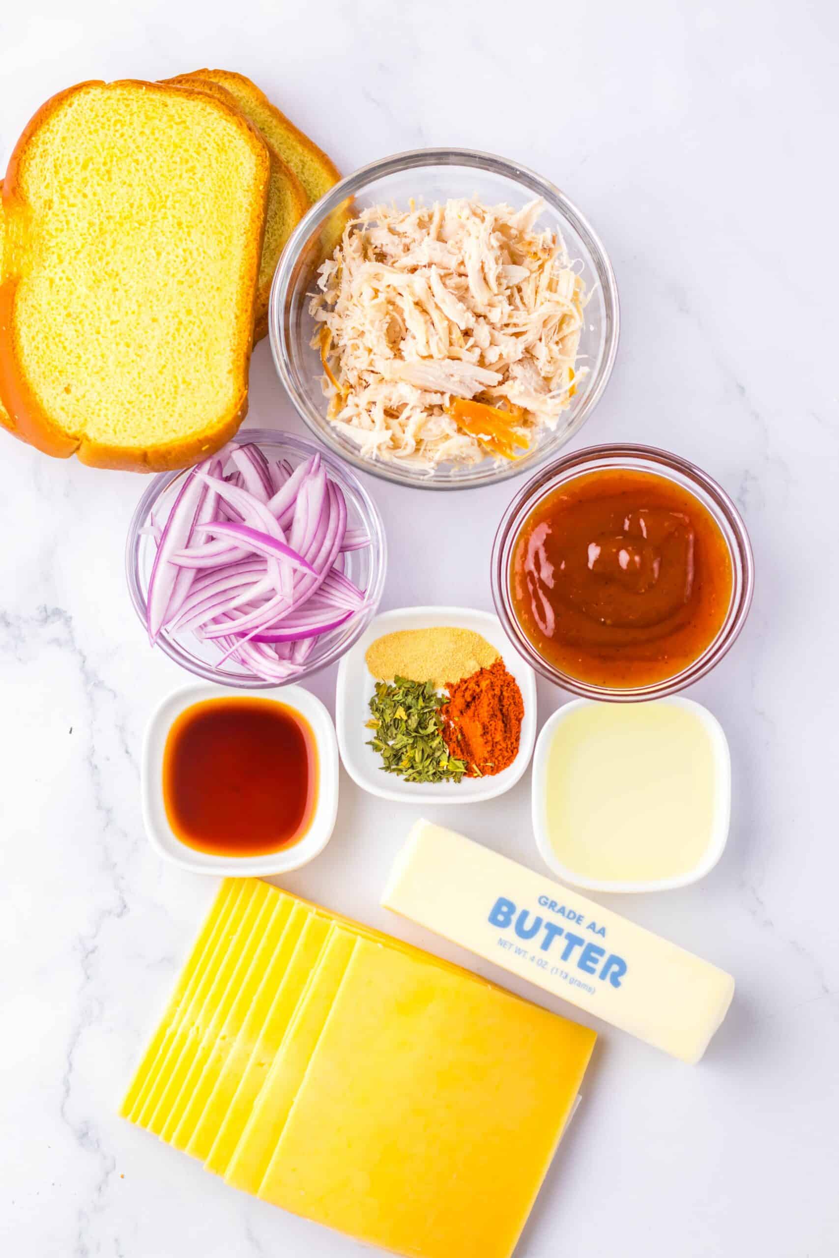 Ingredients arranged on a surface, including two slices of bread, shredded chicken, sliced red onions, barbecue sauce, butter, cheese slices, Worcestershire sauce, and spices.