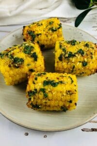 Four pieces of Air Fryer Corn on the cob, coated with herbs and spices, are arranged on a light-colored plate.